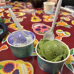 Thai Tea Ice Cream Pint - Two cups of ice cream, one purple and one green, are placed on a table decorated with Day of the Dead-themed stickers, with spoons inserted.