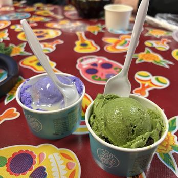 Soft Swerve - Ube Sticky Butter Cake Scoop, Green Thai Tea Oreo Scoop - Two cups of ice cream, one purple and one green, are placed on a table decorated with Day of the Dead-themed stickers, with spoons inserted. - Ice Cream & Frozen Yogurt Near Me - Brooklyn, New York
