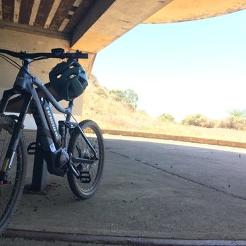 eBikes508 - A black electric mountain bike with a helmet hanging on the handlebar is positioned on a concrete surface beneath a bridge or overpass. The background shows a dry, hilly landscape with sparse vegetation and a clear sky. The bike features a sturdy frame, thick tires, and a visible motor, indicating it is an e-bike suitable for off-road riding. - Bikes Near Me - Plymouth, Massachusetts