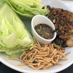 Lettuce Wraps - The image shows a white plate with fresh green lettuce leaves, a small bowl of soy sauce with sesame seeds, crispy chow mein noodles, and a portion of cooked vegetables mixed with ground meat. The dish appears to be lettuce wraps, served with a dipping sauce and crunchy noodles, on a black table. In the background, part of another dish with fried items and lemon slices is visible.