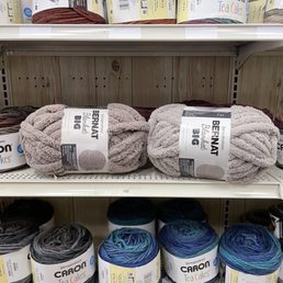 Yarn selection, BOGO 50% off! - Multiple yarn balls are arranged on store shelves in a retail setting. The top shelf features yarn from the Caron Tea Cakes line, with multicolored skeins. The middle shelf displays Bernat Big plush yarn in neutral tones, while the bottom shelf shows Caron Tea Cakes yarn in darker shades, including blue and gray. The shelves are made of wood and metal, with a wooden wall background.