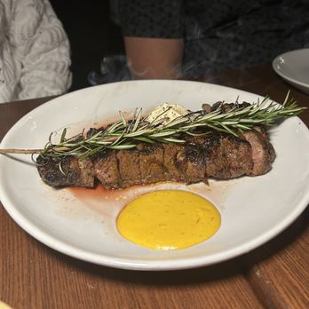 Platea - New York strip - A steak is presented on a white plate, garnished with a sprig of rosemary and accompanied by a yellow sauce, with steam rising from the dish. - Peruvian Near Me - Pinecrest, Florida
