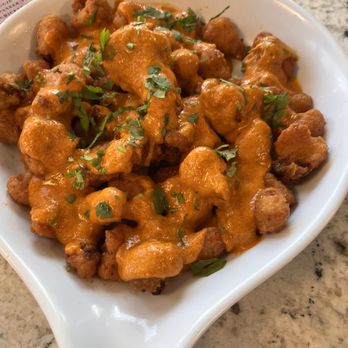 Tikka TapRoom - Cauliflower wings with tikka sauce - Cauliflower wings with tikka sauce are presented in a white, curved bowl. The dish features bite-sized cauliflower pieces coated in a rich, orange-colored tikka sauce, garnished with chopped cilantro. The bowl is placed on a marble countertop, with a partial view of a menu or paper in the background, indicating a dining setting at an Indian fusion restaurant. - Indian Near Me - Lindenhurst, Illinois