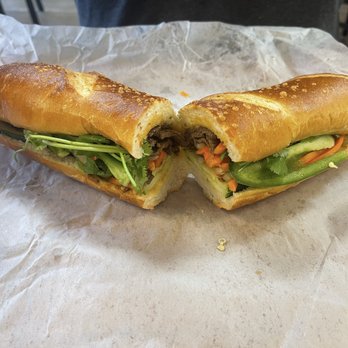 Wrap N Roll - 10. Lemongrass Beef Sandwich - A Vietnamese banh mi sandwich is cut in half, revealing beef, cucumber, carrots, cilantro, and other fresh vegetables inside a toasted baguette. The sandwich is placed on a piece of parchment paper on a wooden surface, with some crumbs visible. The bread has a golden-brown crust, and the filling includes sliced beef and crisp vegetables. - Vietnamese Near Me - Albuquerque, New Mexico