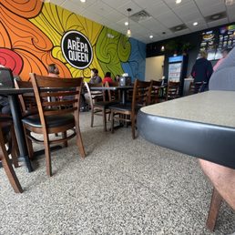 the interior of the restaurant - Inside Arepa Queen, a Venezuelan eatery, with colorful wall art, wooden chairs, tables, and customers dining.