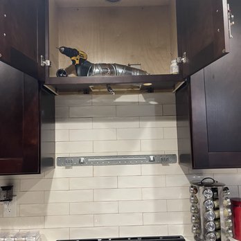Plus58 Remodeling - Removed the microwave hood - A kitchen cabinet is open above a stove, containing a drill, a water bottle, and a small container. The cabinet is part of a kitchen with tiled backsplash and countertop. - General Contractors Near Me