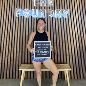 The Foundry - Cheesin - A woman in a black tank top and patterned shorts stands barefoot in front of a wooden wall with a neon sign for The Foundry, holding a sign about her fitness class. - Yoga Near Me - Avondale, Arizona