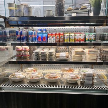 Command Coffee - Selection of grab n go options - The image shows a refrigerated display case filled with bottled beverages, yogurt containers, pre-packaged salads, and desserts. The top shelf has cups, jars, and a plant, while the lower shelves contain food items in plastic containers. A menu is partially visible in the background, indicating a cafe or food service setting. - Coffee & Tea Near Me - Indianapolis, Indiana