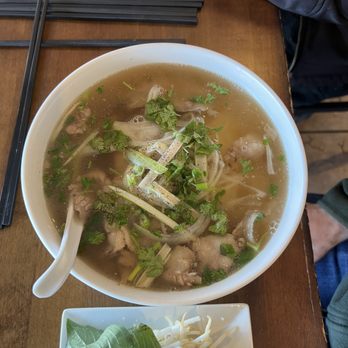 Bamboo by Kulantro - A bowl of Vietnamese pho features clear broth, rice noodles, slices of meat, and garnished with chopped green herbs and onions. A white ceramic spoon rests inside the bowl. Next to the bowl, a small white plate holds fresh basil leaves and bean sprouts. The meal is set on a wooden table, with black chopsticks placed nearby. Part of a person's arm and clothing are visible in the background. - Vietnamese Near Me - Albuquerque, New Mexico