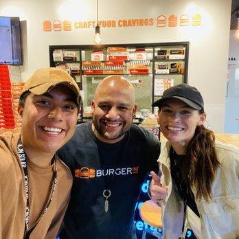 BurgerIM - A group of three people, two women and one man, are taking a selfie inside BurgerIM, a burger restaurant. They are smiling and appear happy, with the woman on the left wearing a tan cap and brown shirt, the man in the middle wearing a black BurgerIM shirt, and the woman on the right wearing a black cap and light-colored jacket. The background features a menu shelf with burger boxes, a TV screen, and warm lighting fixtures. The setting is casual and modern, with a focus on the group and the restaurant's interior. - Burgers Near Me - Brambleton, Virginia