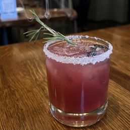 A beverage served in a short, clear glass with a salted rim. The drink is pinkish-red and contains ice cubes. A sprig of rosemary is placed as a garnish on top of the drink. The glass is on a wooden table, with a blurred background suggesting an indoor setting.