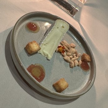 JORY - A ceramic plate holds a rectangular piece of cheese, two small crackers, a handful of nuts, and dollops of sauces, arranged on a restaurant table with a glass of water nearby. The setting suggests a fine dining environment with a white tablecloth. - New American Near Me - Newberg, Oregon