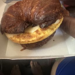 Burnt Bacon Croissant - A croissant sandwich with melted cheese is held by a person's hand, with a burnt bacon croissant placed on top. The sandwich appears to be inside a paper-lined tray, with a red cup and other items visible in the background. The person's feet are visible at the bottom of the image, and the setting appears to be a casual dining or fast-food environment.