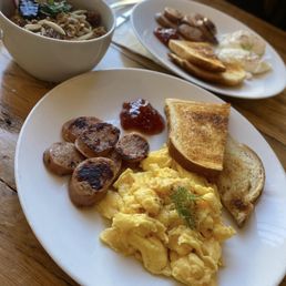 A breakfast plate includes scrambled eggs garnished with a sprig of herbs, grilled sausage slices, two pieces of toasted bread, and a small serving of jam. In the background, there is a bowl of noodle soup with various toppings, placed on a wooden table. The setting appears to be a casual dining environment.