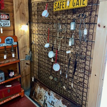 Ruby’s Mercantile - The image shows a wire grid display mounted on a wooden wall, featuring vintage kitchen utensils such as small pans, scissors, and ladles, each with price tags. A yellow sign reading 'SAFETY GATE' is positioned at the top of the grid. To the left, part of a shelving unit with jars and a red toolbox is visible. The setting appears to be an antique or thrift store with a rustic, nostalgic atmosphere. - Antiques Near Me - Westcliffe, Colorado