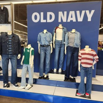 Old Navy - Mannequins in casual denim outfits and t-shirts are positioned on a platform in front of a large blue sign with white text reading 'OLD NAVY'. The display includes various styles of jeans, jackets, and shirts, with some mannequins styled with layered clothing. The store interior with clothing racks and lighting is visible in the background. - Men's Clothing Near Me - Davie, Florida