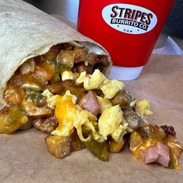 Breakfast Burrito - A breakfast burrito filled with meat, cheese, and vegetables is partially unwrapped on a paper surface, with a Stripes Burrito Co. cup in the background.