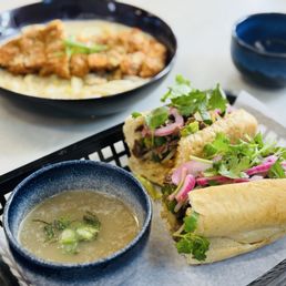 Creamy Udon - Two Vietnamese banh mi sandwiches filled with herbs, pickled vegetables, and meat are placed on a black tray lined with paper. A small bowl of broth with green onions is also on the tray. In the background, there is a bowl and a larger dish with a noodle and meat dish, likely pho dip. The setting appears to be a casual dining environment.