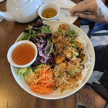 Bamboo by Kulantro - A large white plate features grilled shrimp topped with fried shallots and chopped green onions, served alongside rice noodles and a variety of fresh vegetables including cucumber slices, shredded carrot, and purple cabbage. Two small bowls contain dipping sauces, one with a clear, orange-colored sauce and the other with a darker, amber-colored tea. A white teapot and a cup of tea are on the wooden table, with a person's hand holding a fork nearby. The setting appears to be a casual dining environment, likely a Vietnamese restaurant. - Vietnamese Near Me - Albuquerque, New Mexico