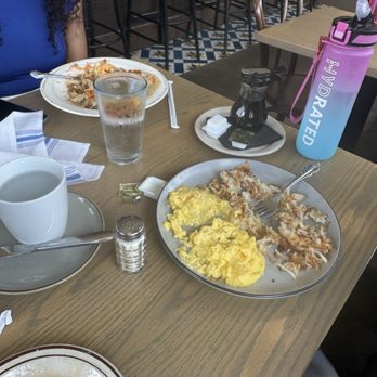 The Harvest Room - Hash and Eggs - A breakfast plate with scrambled eggs and hash browns is on a wooden table, alongside a coffee mug, a glass of water, and a pink water bottle. - New American Near Me - Munster, Indiana