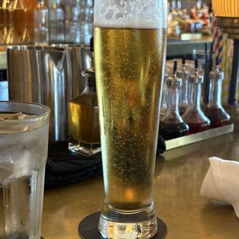 Goodwin’s - Beer - A tall, slender glass of beer with bubbles and foam on top is placed on a bar counter, with bar equipment and bottles in the background. - Cocktail Bars Near Me - Dallas, Texas