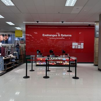 Target - A retail store's exchanges and returns counter area features a large red wall with white text indicating 'Exchanges & Returns' and its Spanish translation. There are multiple checkout stations with cash registers and card readers, separated by black stanchions with red and green belts. To the left, clothing displays and mannequins are visible, indicating the store sells apparel. The floor is light-colored tile, and the ceiling has fluorescent lighting panels. - Department Stores Near Me - Hackensack, New Jersey