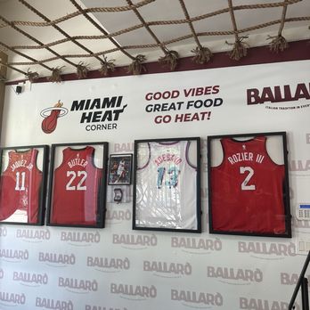 Ballaro - The image shows four framed Miami Heat basketball jerseys hung on a wall inside a restaurant or sports-themed establishment. The jerseys are displayed in black frames, with the players' names and numbers visible. The wall features Miami Heat branding and motivational phrases such as "Good Vibes," "Great Food," and "Go Heat!" The ceiling has decorative rope accents, and there are small potted plants and a tissue box on a table in the foreground. - Italian Near Me - Miami, Florida