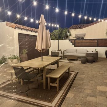 Illuminating Team - A nighttime outdoor patio features string lights overhead, a wooden dining table with benches, a seating area with sofas, and potted plants along the walls. - Lighting Fixtures & Equipment Near Me