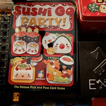 Solid State - The image shows a box for the card game 'Sushi Go Party!' with colorful, cartoon-style illustrations of sushi characters, including nigiri, rolls, and other Japanese cuisine items. The box indicates it is suitable for ages 8 and up and supports 2-8 players. The game is described as a deluxe pick and pass card game, with branding from Gamewright. The box is placed among other board games on a shelf. - Bars Near Me - Woodside, New York