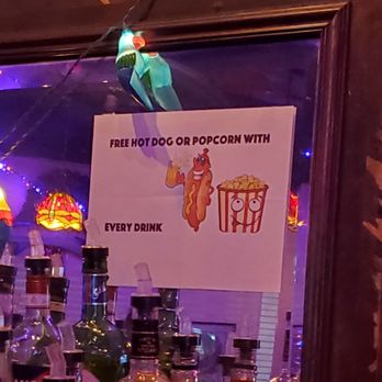 Myrtle Pub - a sign on a bar counter - A sign in a bar offers a free hot dog or popcorn with every drink purchase, featuring cartoon images of a hot dog and popcorn. - Bars Near Me - Queens, New York