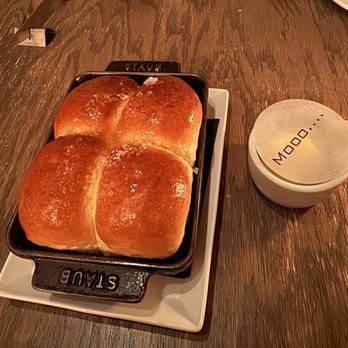 Mooo.... Beacon Hill - Three bread rolls with a shiny, golden-brown crust are in a black cast iron skillet on a white plate, set on a wooden table. - Steakhouses Near Me - Boston, Massachusetts