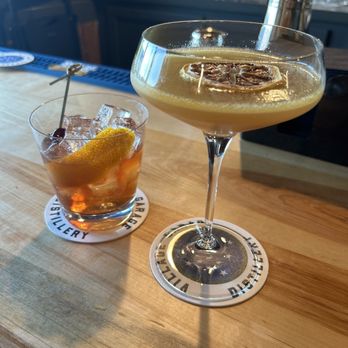 The Trailhead - Old Fashioned & Fig-Ure It Out - Two cocktails are displayed on a wooden bar counter. The first is in a short glass filled with ice, a lemon twist, and a dark amber liquid. The second is in a coupe glass with a pale yellow drink, garnished with a lemon slice and a large ice cube. Both drinks are placed on branded coasters, and the background shows a bar setting with glasses and bar equipment. - Bars Near Me - Peru, Vermont
