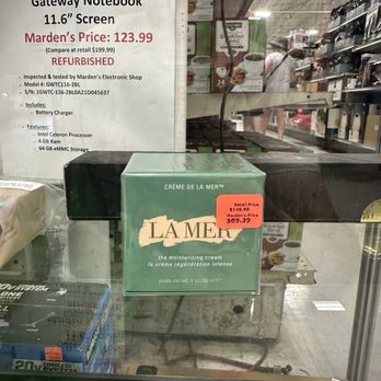 Marden's - Creme de la Mer by La Mer. - A green box of La Mer moisturizing cream, labeled 'Crème de la Mer,' is positioned behind a glass display case in a retail store. The box has a red price sticker indicating a sale price of $89.39, down from $148.98. In the background, store shelves and a sign advertising a refurbished Marden's Gateway Notebook with a 11.6-inch screen are visible, along with a person shopping. - Discount Store Near Me - Biddeford, Maine