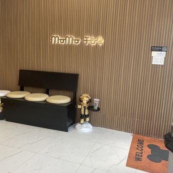 MoMo Tea - Inside - The image shows the interior of a business with a wooden wall, illuminated sign, black bench with cushions, a gold astronaut statue, and a welcome mat. - Bubble Tea Near Me - Bellmore, New York