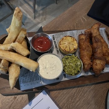John Martin's - A meal featuring three pieces of battered fried fish fillets, a lemon half, French fries, and three small containers of condiments (ketchup, tartar sauce, and relish) on a wooden serving tray lined with paper. The tray is placed on a wooden table in a restaurant setting. - Irish Near Me - Miami, Florida