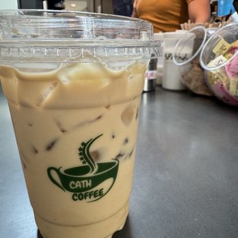 Cath Coffee - Almond latte - A plastic cup filled with iced almond latte, featuring the Cath Coffee logo, is placed on a counter at a coffee shop. - Coffee & Tea Near Me - Indianapolis, Indiana