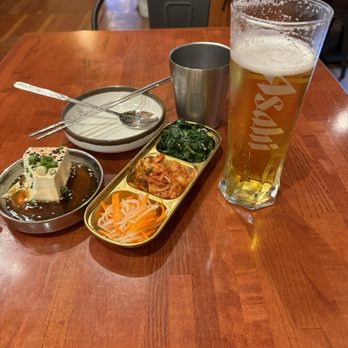 Tiger Mama - Starter... I anticipate greatness - A wooden table holds a tall glass of beer, a metal cup, a bowl with a spoon, and a tray with side dishes including kimchi, spinach, and pickled vegetables. - Korean Near Me - Eugene, Oregon