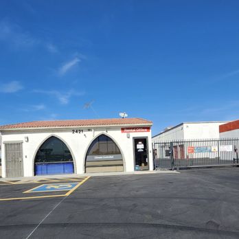 Public Storage - The Office - A small, white, single-story building features two large arched windows and a door with a sign indicating it is a rental office. The building has a red-tiled roof and is located in a paved parking lot with a designated handicap parking space. The sky above is clear and blue, with a few wispy clouds. Surrounding the building are other structures and a fence, indicating a commercial or industrial area. - Self Storage Near Me - Phoenix, Arizona
