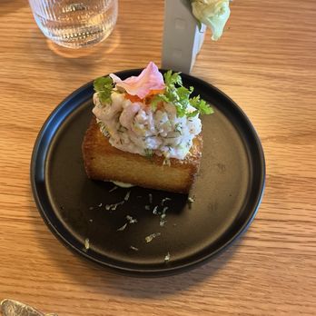 Domek - Shrimp toast - A serving of shrimp salad, topped with herbs and flower petals, is presented on a toasted bread slice on a black plate. The plate is on a wooden table, accompanied by a small white vase with white roses, a glass of water, and a knife. The setting suggests a dining experience at a restaurant or cafe. - Modern European Near Me - Eugene, Oregon