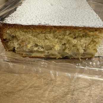 Brooklyn Bread House - Apple cake - A rectangular slice of apple cake with a dusting of powdered sugar on top is displayed on a clear plastic tray. The cake has a moist, crumbly texture with visible apple pieces embedded within. It is set on a wooden surface, with a blurred background indicating an indoor setting, likely a bakery or cafe. The cake appears freshly baked and ready for serving. - Bakeries Near Me - Brooklyn, New York
