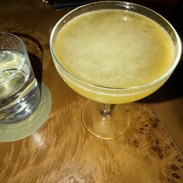 A yellow cocktail in a coupe glass is positioned on a wooden table, with a glass of water to its left. The table has a natural wood grain finish, and a black purse is visible in the background. The setting appears to be in a dimly lit environment, possibly a bar or lounge.