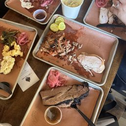 Several trays with remnants of barbecue meats, pickles, and sides are on a wooden table, with cups of sides and sauces.