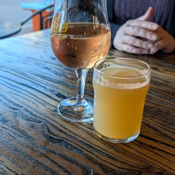 Fass Brewing - 4 ounce pour and wine - Two drinks, one rosé wine and one pale beer, are placed on a wooden table inside a restaurant or bar, with a person sitting nearby. - Breweries Near Me - Conroe, Texas