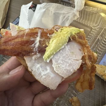 Corner Bakery Cafe - Small single piece of avacado - A sandwich featuring bacon, avocado, turkey, and lettuce is held in a person's hand. The sandwich is assembled with various ingredients visible, including crispy bacon, sliced avocado, turkey, and lettuce, on bread. The background shows a paper wrapper and a plastic container, indicating a takeout or casual dining setting. - Cafes Near Me - El Paso, Texas