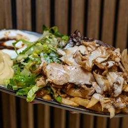 Mix Shawarma Plate - A large, oval-shaped plate contains a serving of mixed shawarma, including sliced meat, topped with sauce. Accompanying the shawarma are fresh chopped lettuce and other greens, along with dollops of hummus and mayonnaise, and a drizzle of dark sauce. The plate is held by a person’s hand, with a wooden slat background behind it.