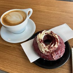 Oat milk latte and red velvet doughnut - A white ceramic cup of coffee with foam sits on a saucer, next to a red velvet doughnut with white icing and red sprinkles on a black plate.