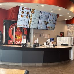 A restaurant counter features digital menu screens displaying food options, with promotional posters on the wall and a cash register.