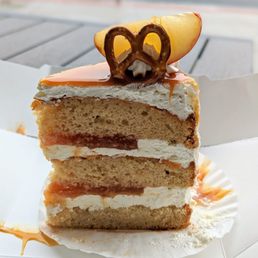 Apple Cake - A layered cake with cream, caramel, and apple toppings, decorated with a pretzel and apple slice, served on a white paper tray.