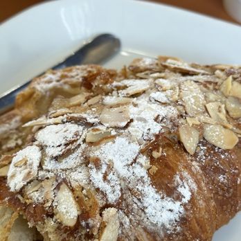 Cafe Barista - Suffield - Almond Croissant - A detailed view of an almond croissant topped with sliced almonds and powdered sugar, served on a white plate with a metal utensil nearby. - Coffee & Tea Near Me - Suffield, Connecticut