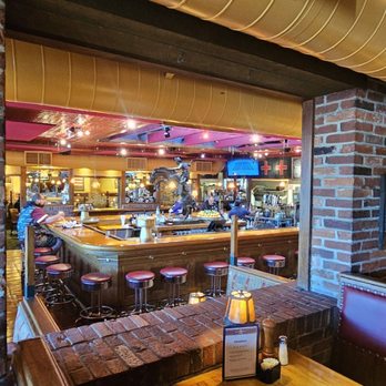 Silverado - The image shows the interior of a restaurant with a bar area in the background, surrounded by brick walls and booths with red upholstery. The ceiling has exposed ductwork and decorative lighting. Patrons are seated at the bar and tables, with some staff visible behind the bar. The setting appears to be a casual dining space with a warm, rustic atmosphere. - American Near Me - Annandale, Virginia